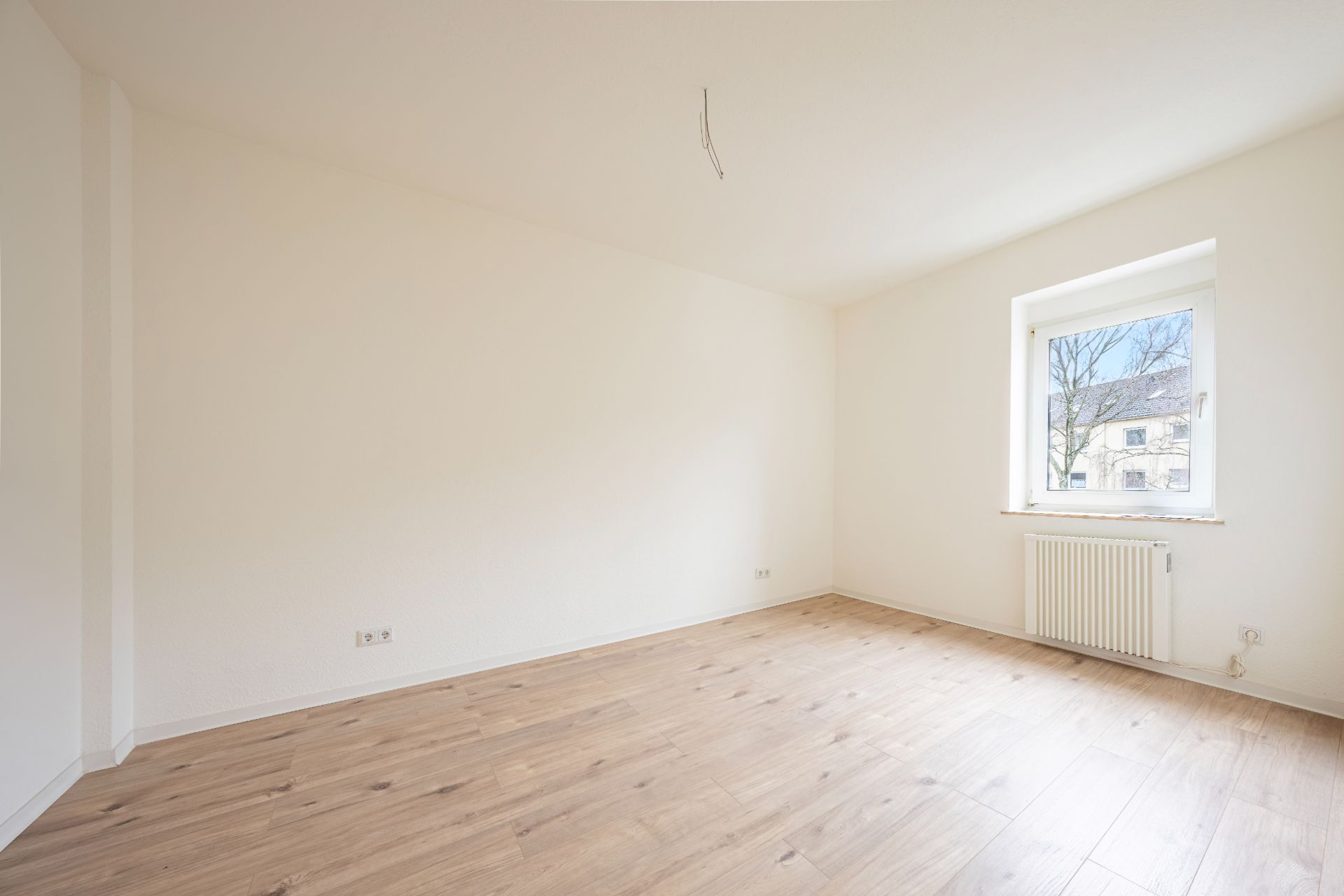 Helles, leeres Zimmer mit Holzfußboden, weißer Wand und einem Fenster mit Blick nach draußen.