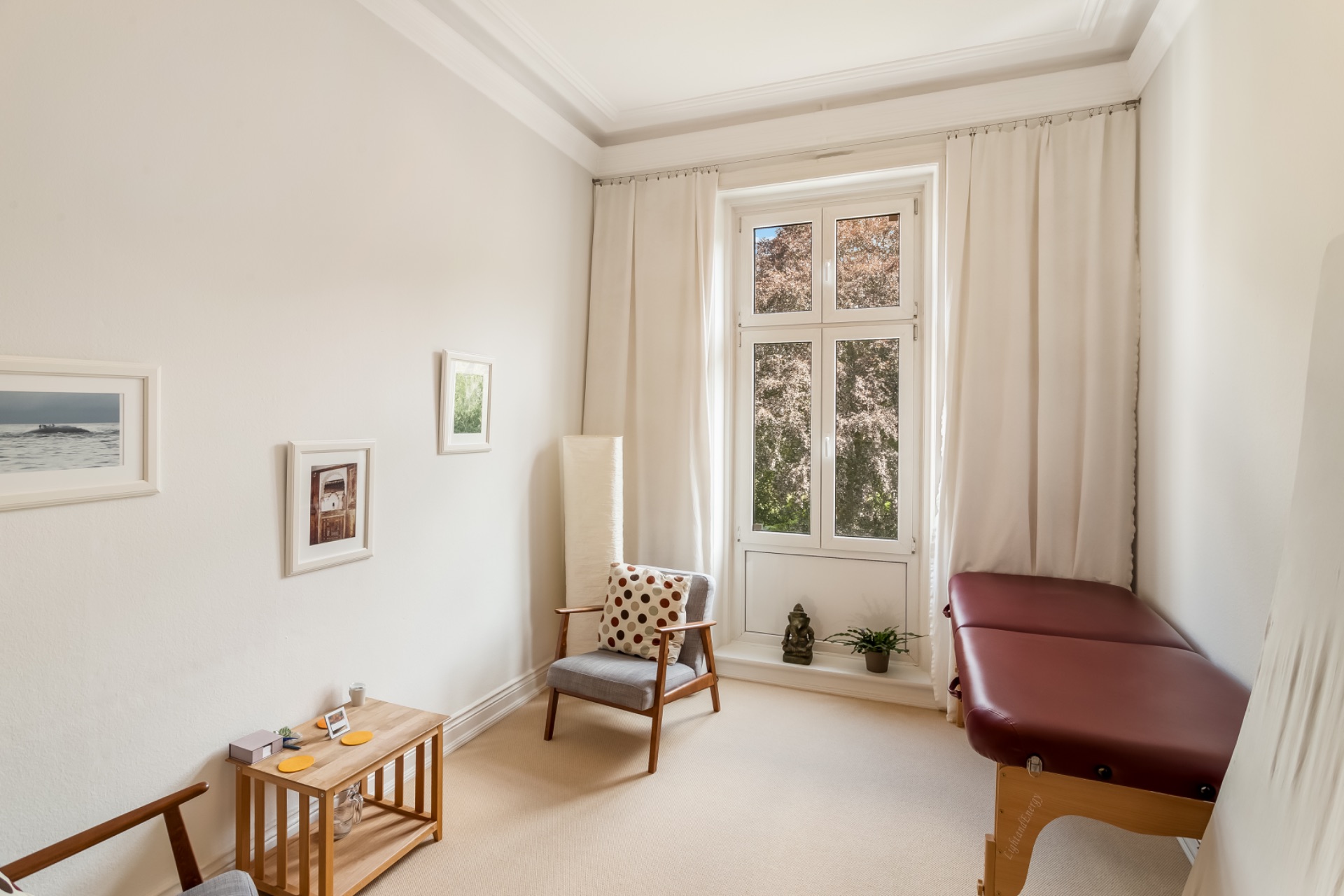 Heller Raum mit Sessel, Bilder an der Wand, Massageliege und großem Fenster mit Vorhängen.