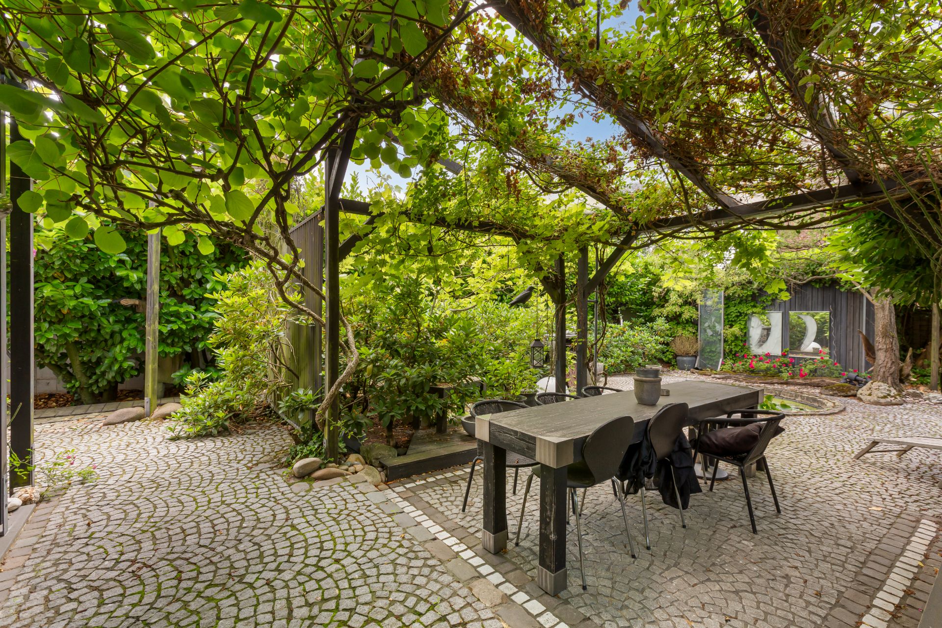 Terrasse mit Tisch und Stühlen unter einer bewachsenen Pergola in einem grünen Garten.