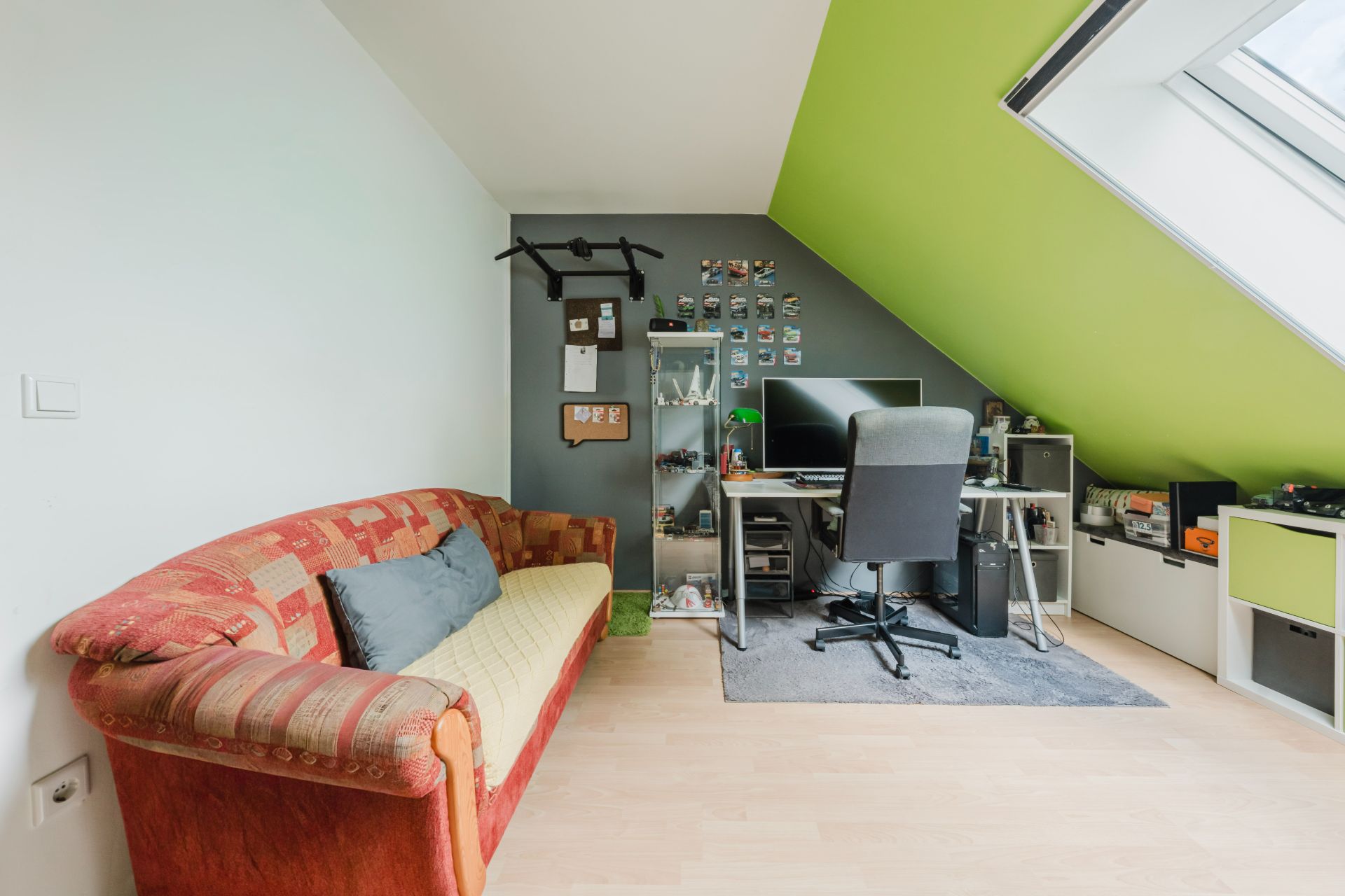 Wohnzimmer mit rotem Sofa, Schreibtisch und Computer unter einer grünen Dachschräge.