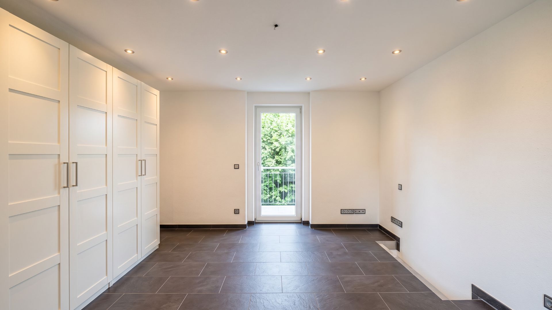 Heller Raum mit Einbauschränken, Fliesenboden und Tür zum Balkon.