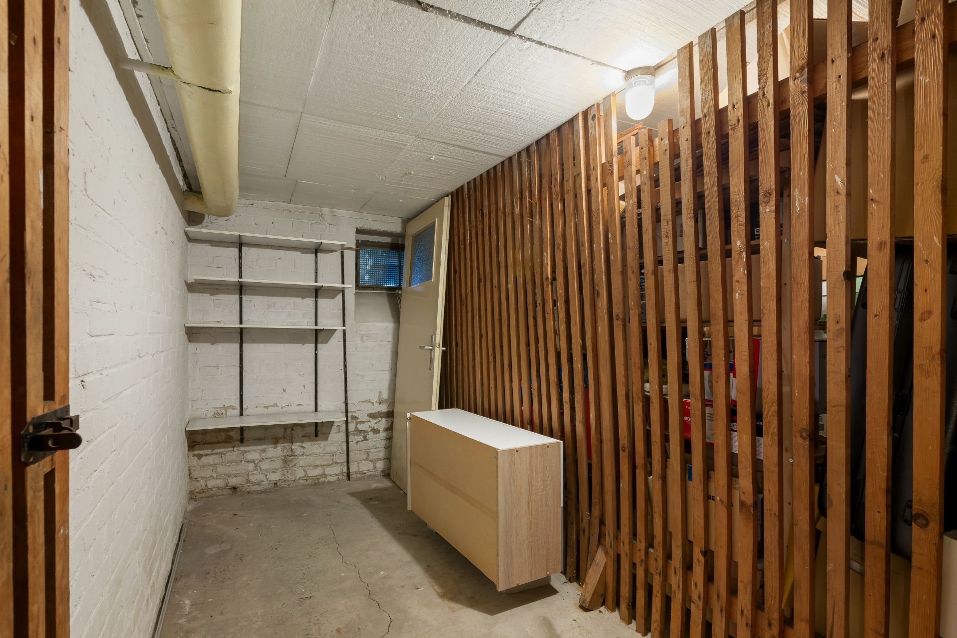 Kellerraum mit Holzgitterwand, Regal, Kommode und weiß gestrichenen Wänden.