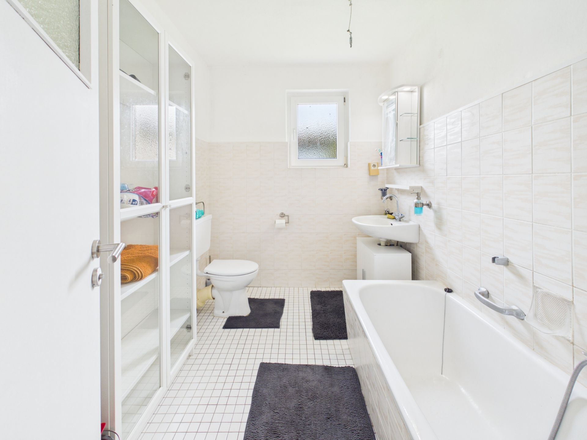 Helles Badezimmer mit Badewanne, Toilette, Waschbecken, Regalen und Fenster.