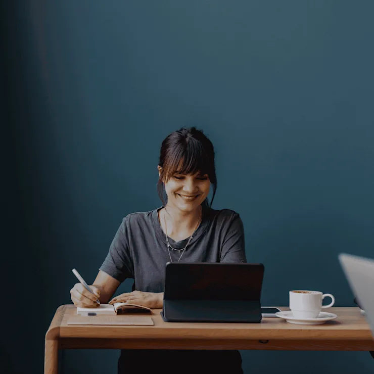 Frau sitzt an einem Holztisch, schreibt und arbeitet an einem Tablet, daneben eine Tasse Kaffee.
