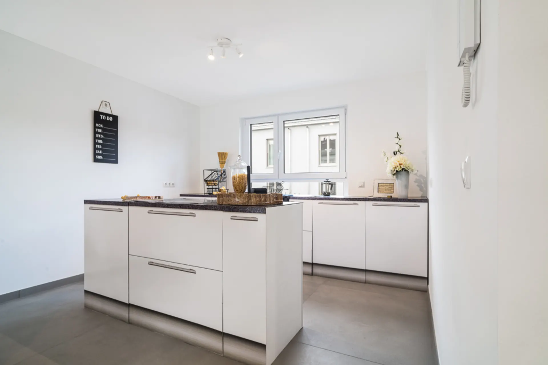 Moderne weiße Küche mit Kücheninsel und großen Fenstern, Dekoration und To-Do-Tafel an der Wand.