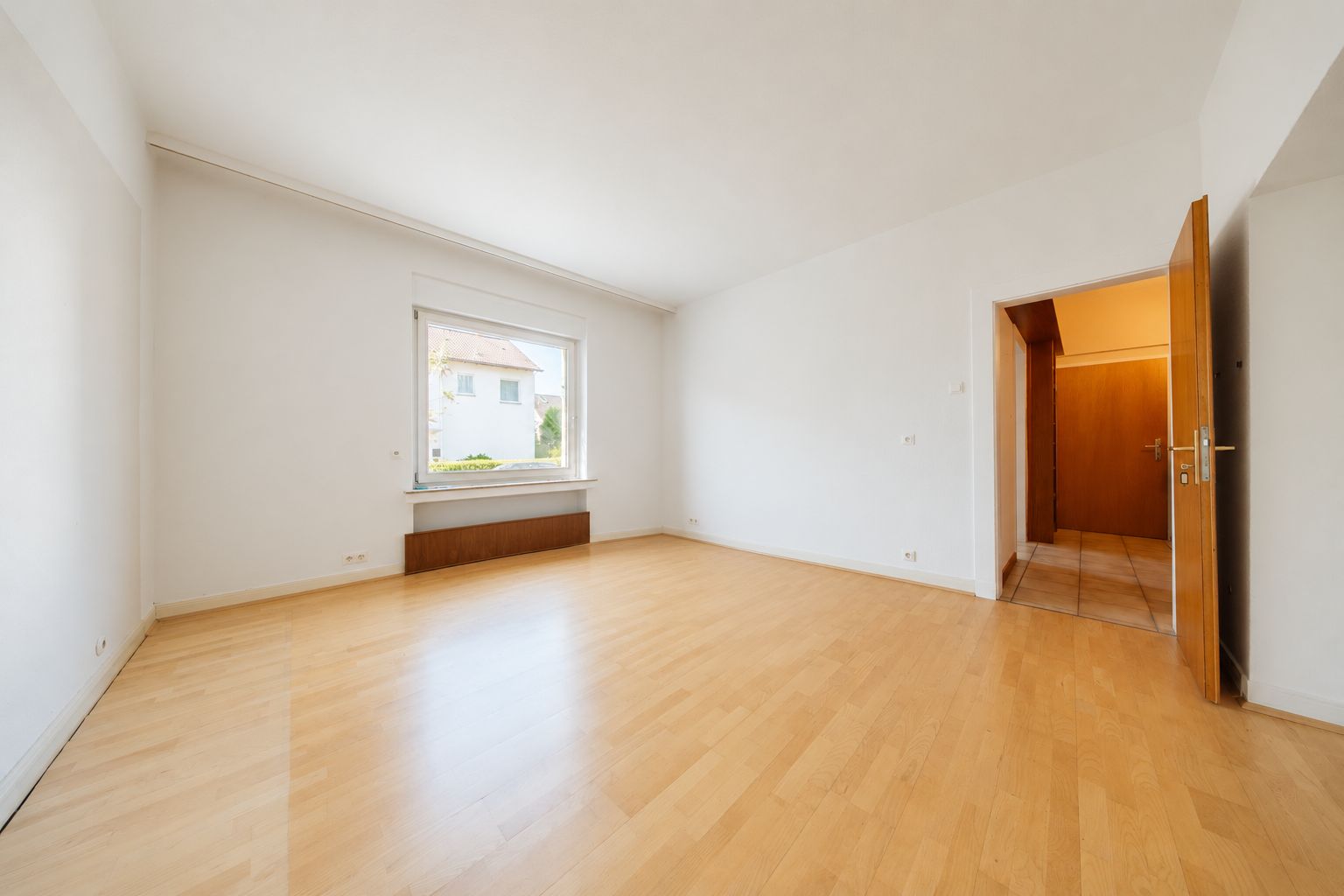 Heller, leerer Raum mit Holzfußboden, großem Fenster und offen stehender Tür.