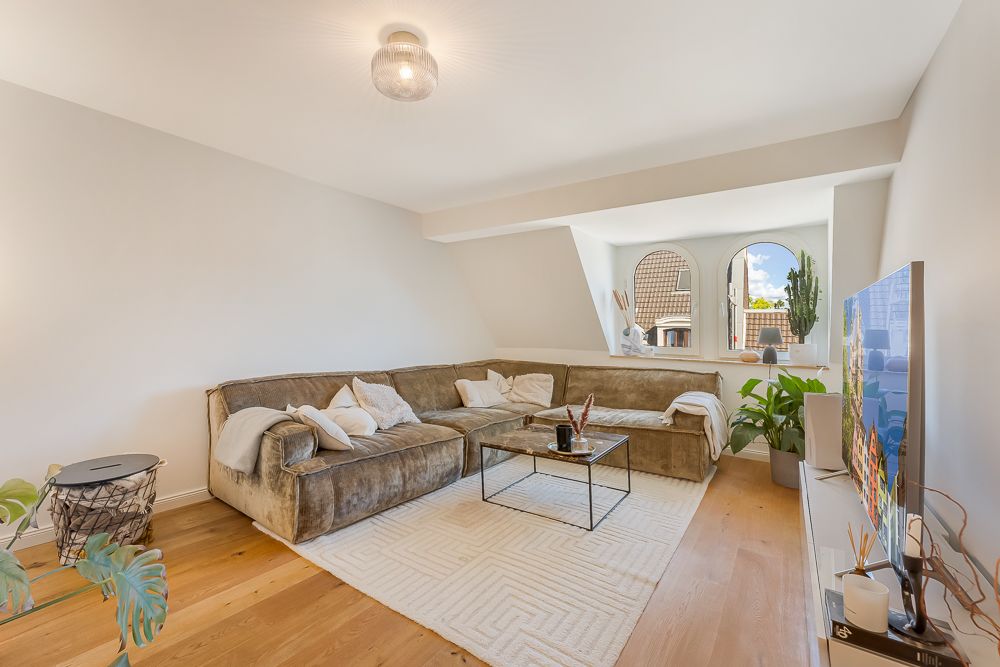 Wohnzimmer mit großem Ecksofa, Glastisch, Fernseher, Pflanzen und zwei Bogenfenstern.