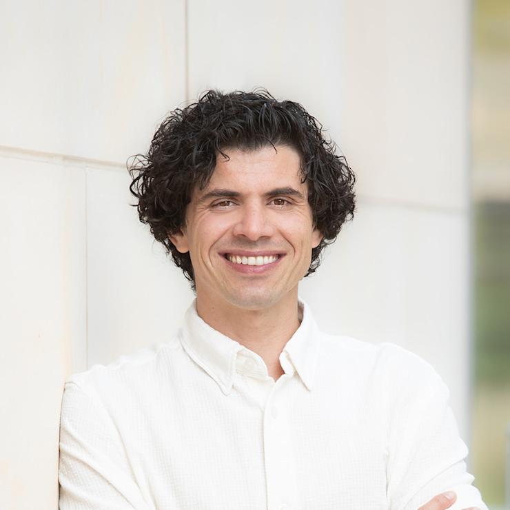 Ein Mann mit lockigen Haaren und hellblauem Hemd steht mit verschränkten Armen vor einer weißen Wand.