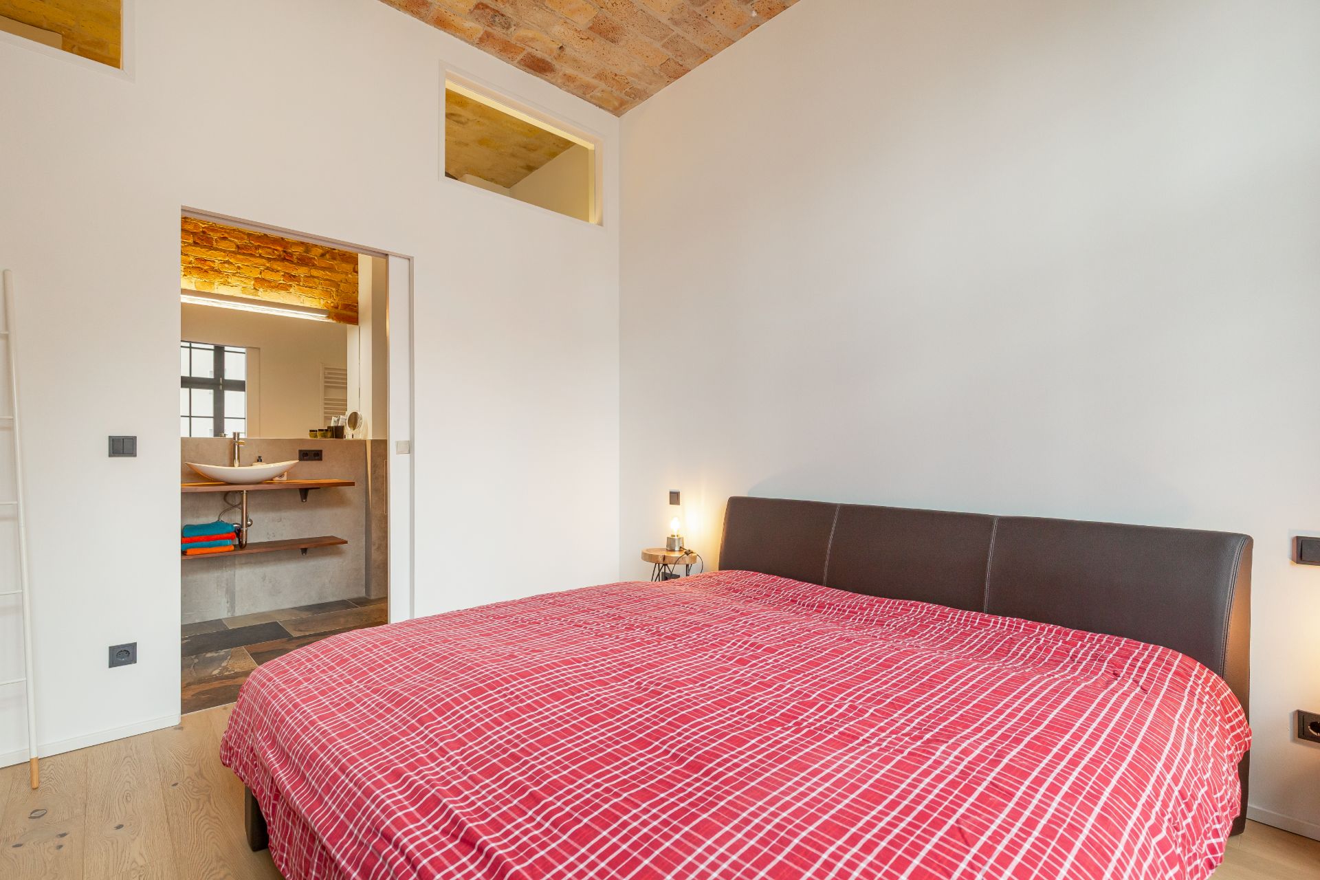 Schlafzimmer mit Doppelbett und roter Bettdecke, Blick ins Badezimmer.