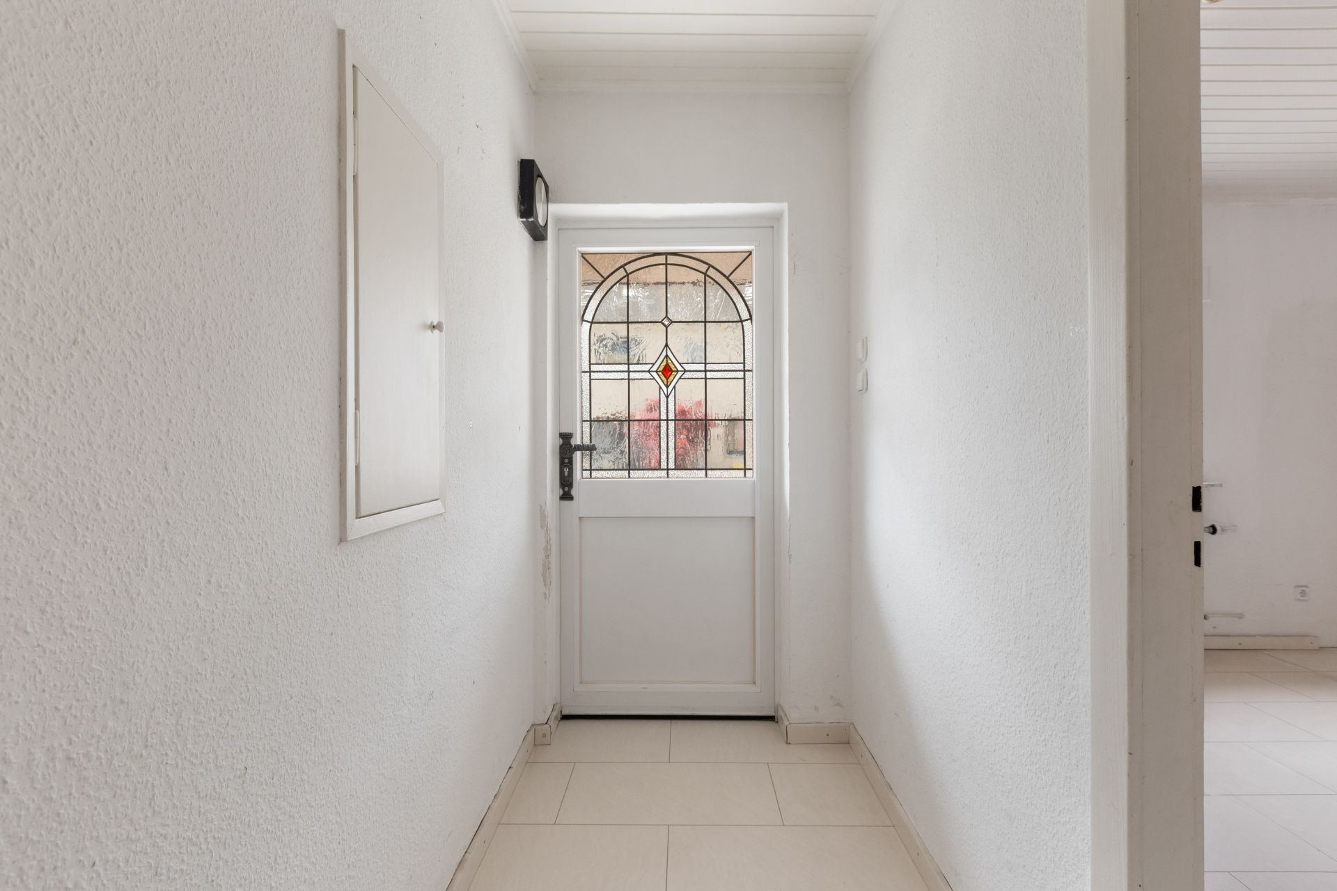Heller Flur mit weißer Tür und buntem Glasfenster, Fliesenboden, weiße Wände.