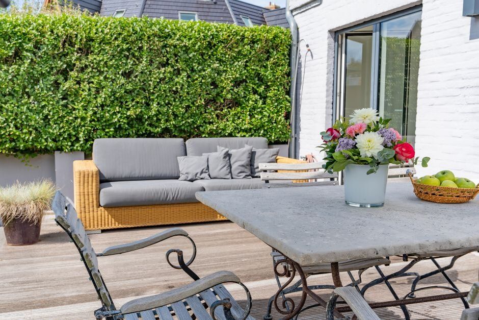 Terrasse mit Tisch, Stühlen, Sofa, Blumenstrauß und Schale mit Äpfeln, vor grüner Hecke.