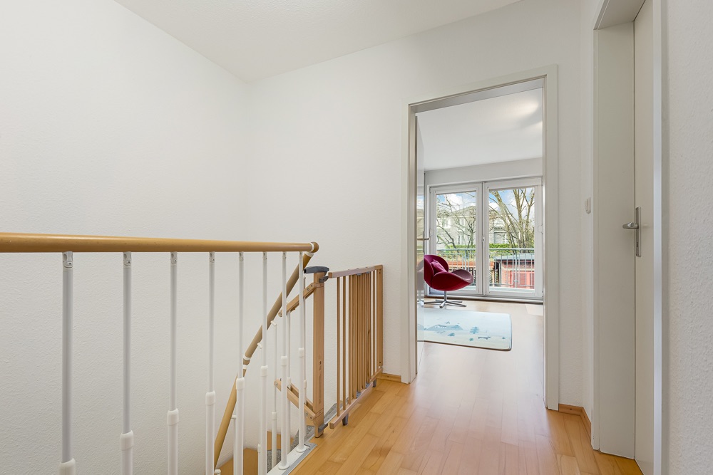 Heller Flur mit Holzboden, Treppengeländer und Blick in ein Zimmer mit rotem Stuhl und großem Fenster.