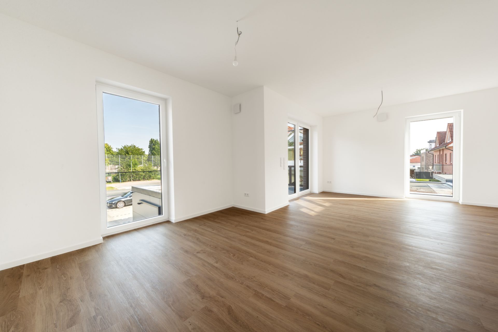 Helles, leeres Wohnzimmer mit Holzfußboden und großen Fenstern.
