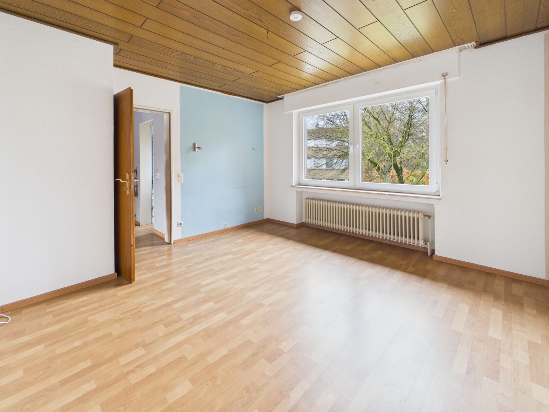 Leeres Zimmer mit Holzdecke, Laminatboden, großem Fenster und Heizkörper unter dem Fenster.
