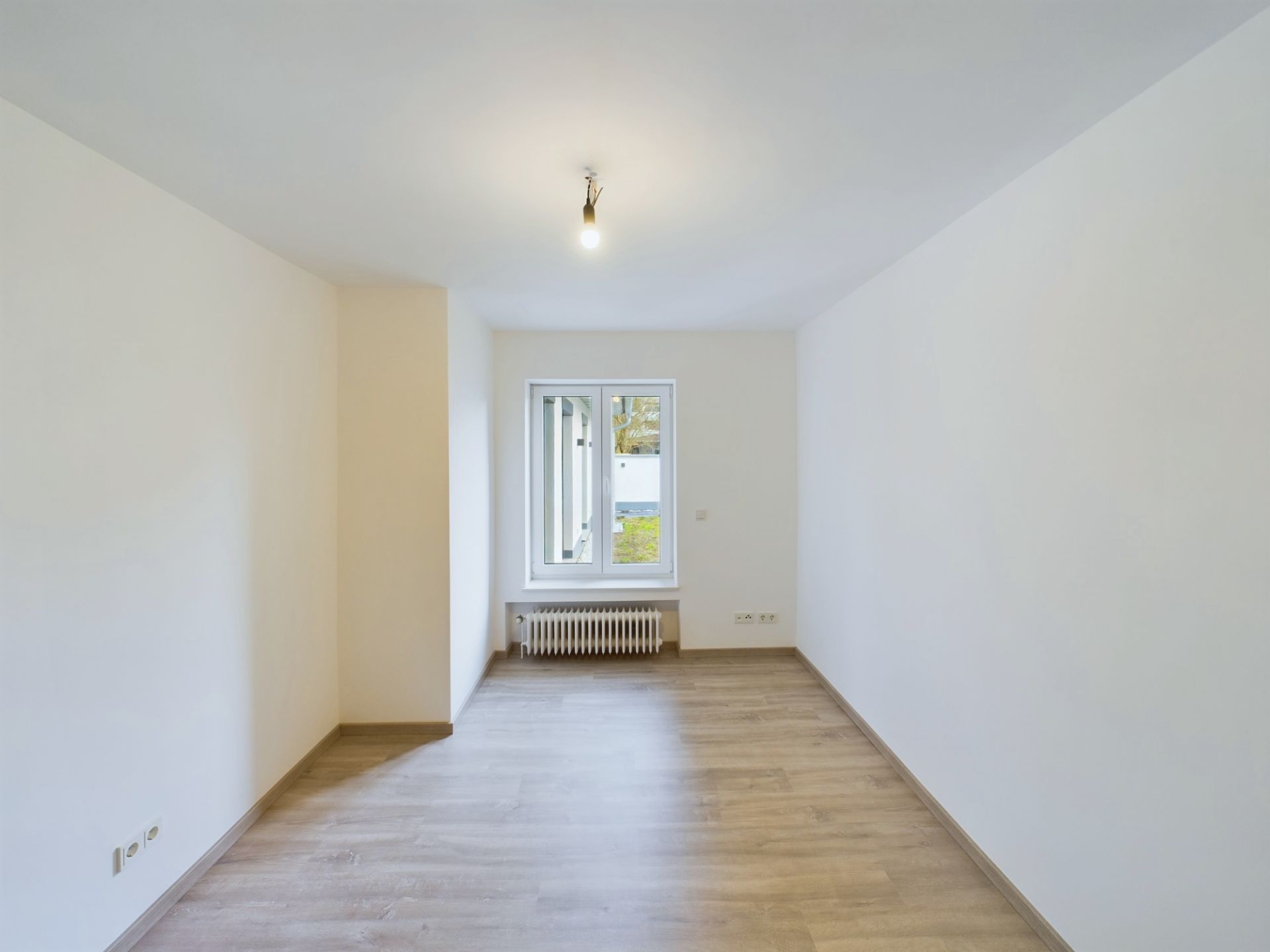 Leerer Raum mit hellem Holzfußboden, weißem Fenster und Heizkörper unter dem Fenster.