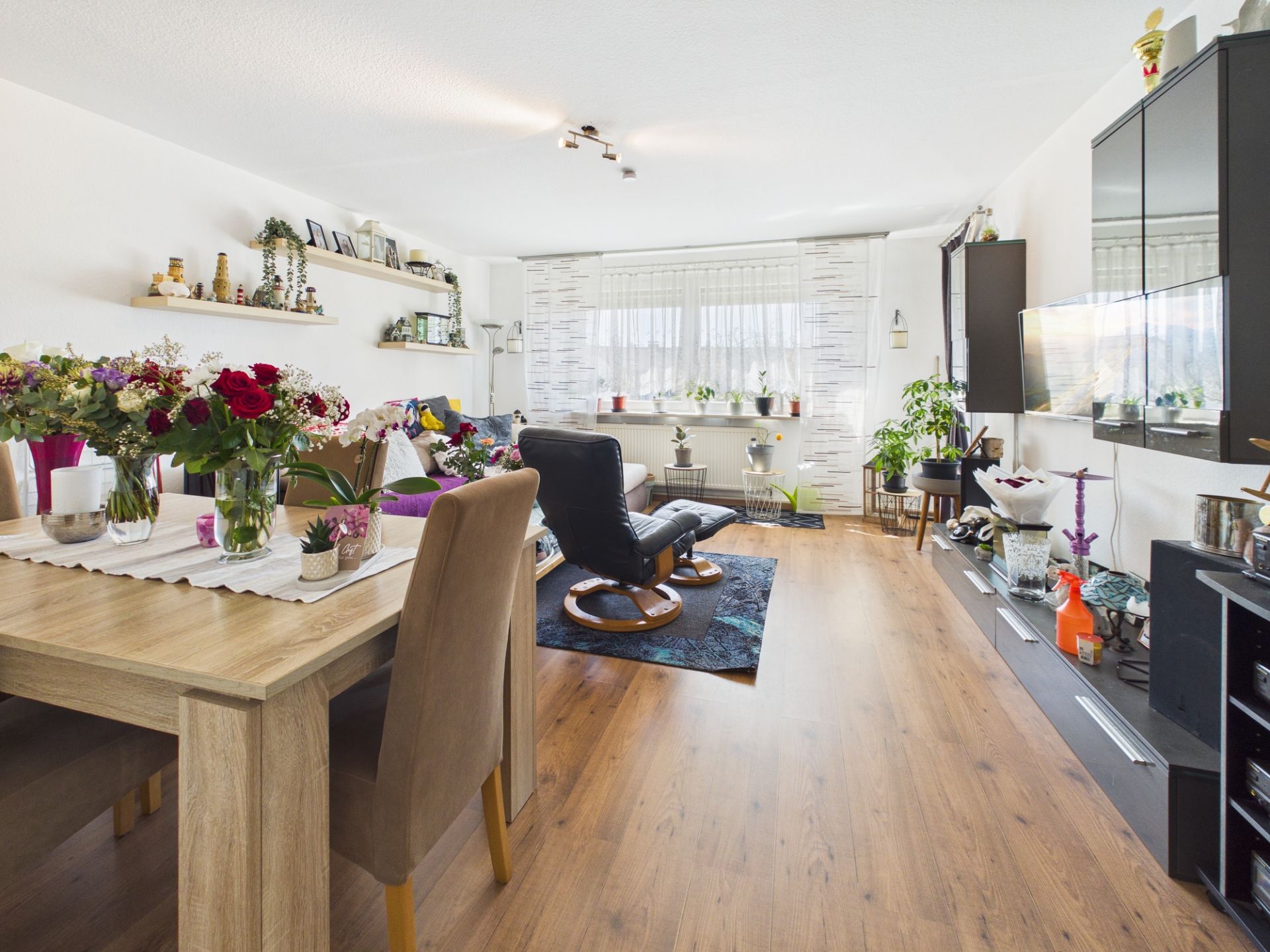 Helles Wohnzimmer mit Esstisch, Stühlen, Blumen und Regal, große Fenster mit Pflanzen.