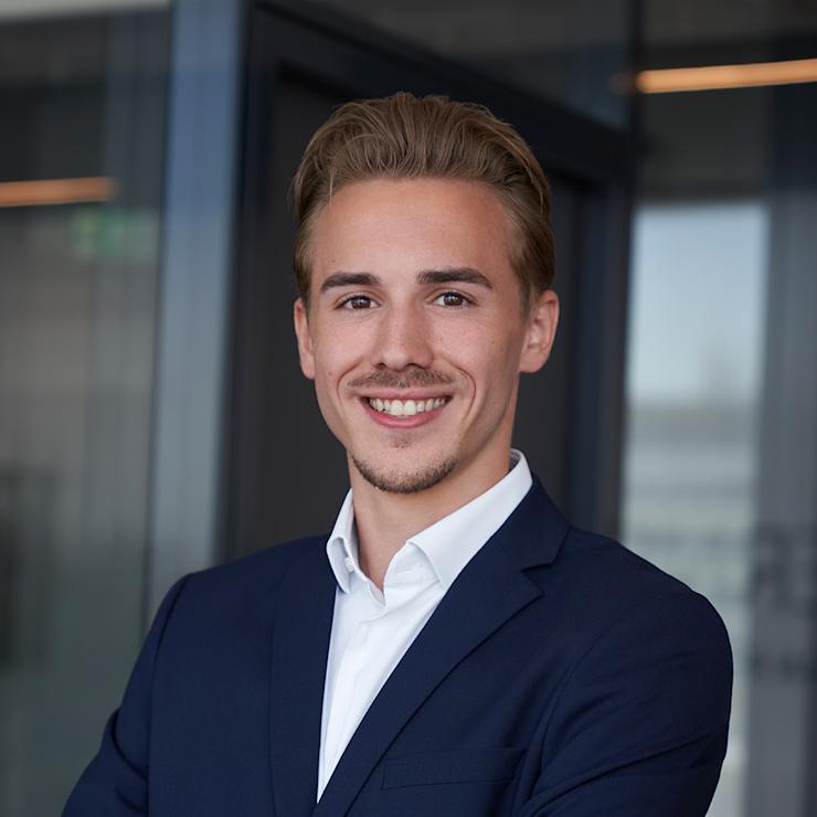 Junger Mann im Anzug steht mit verschränkten Armen vor Glaswand.