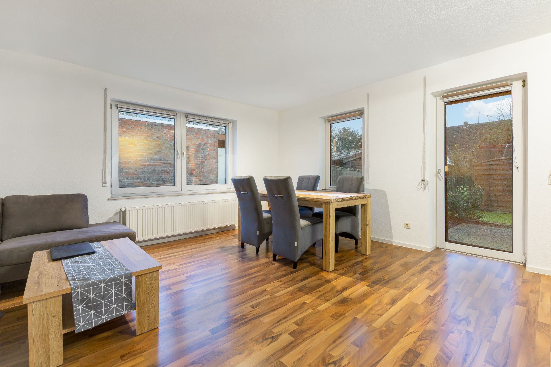 Helles Wohnzimmer mit Holzboden, Esstisch, vier Stühlen, Sofa und Couchtisch, Zugang zum Garten.
