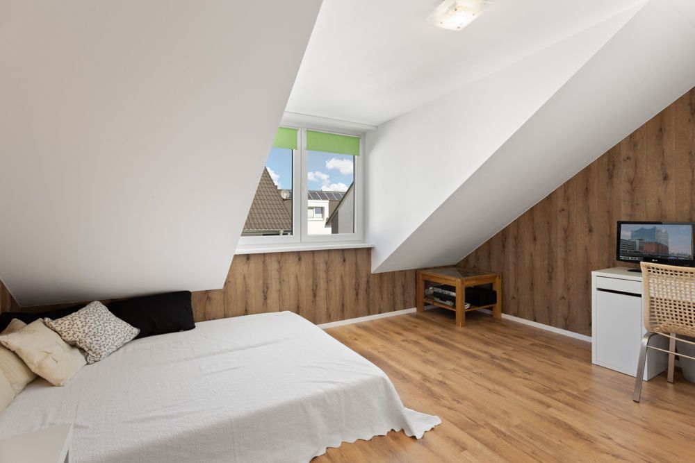 Dachzimmer mit Bett, Holzfußboden, Schreibtisch und großem Fenster.