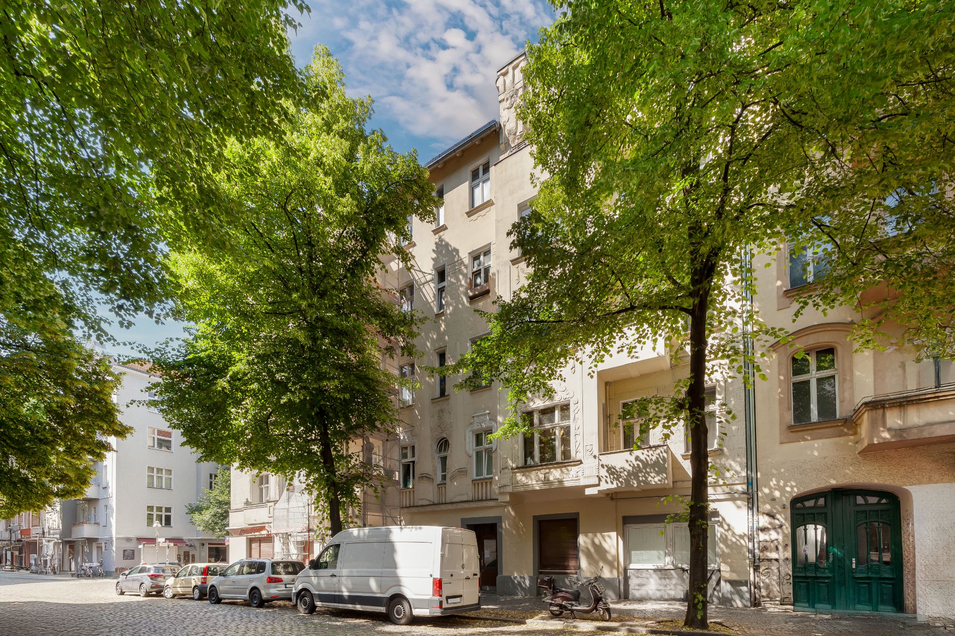 Wohnhaus mit mehreren Stockwerken und Balkonen, vor dem Bäume und parkende Autos stehen.