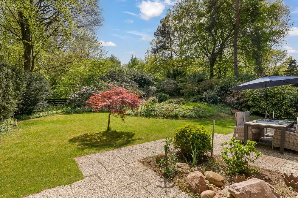 Terrasse mit Esstisch, Sonnenschirm und Blick auf Garten mit kleinem Baum und viel Grün.