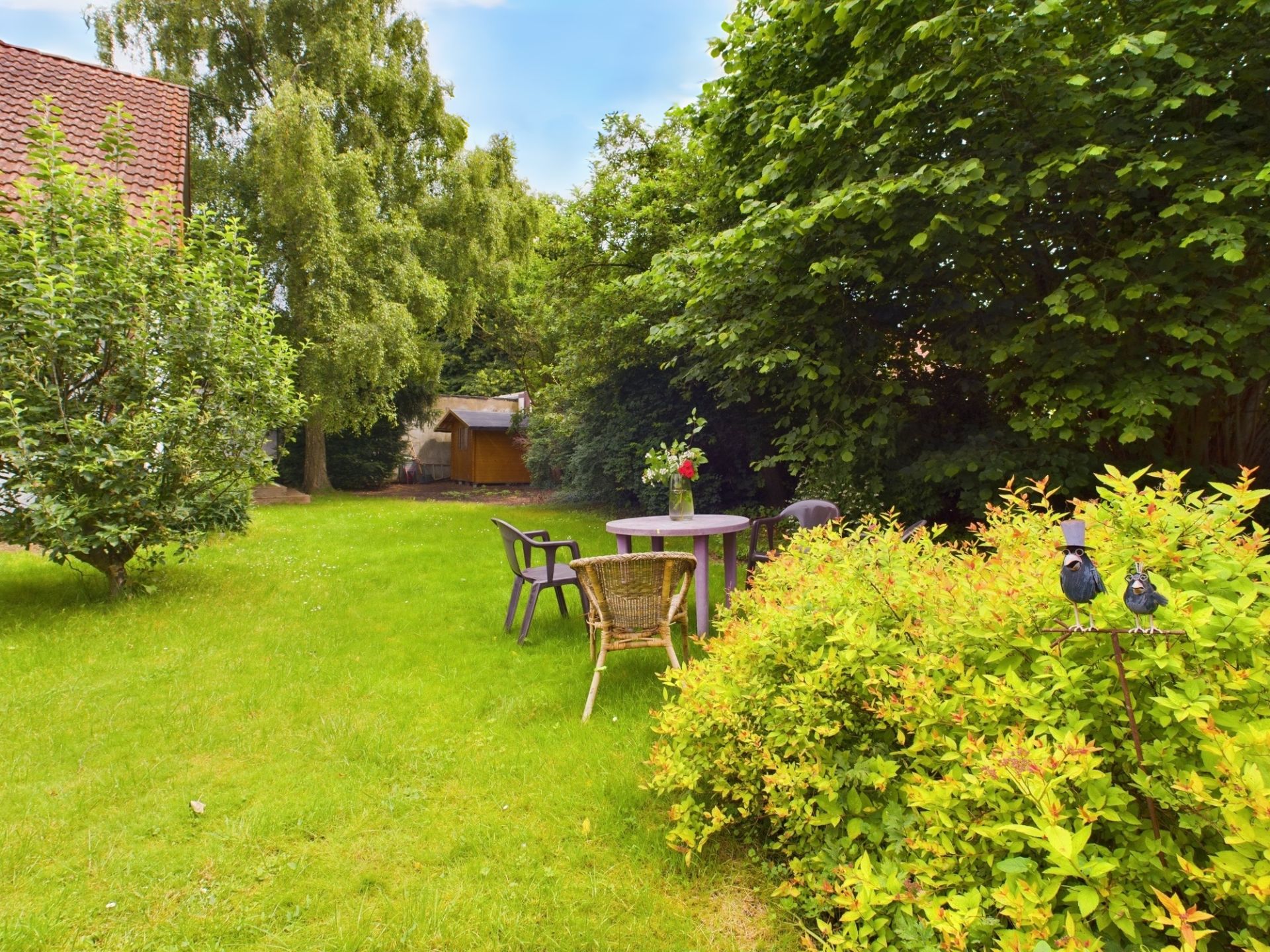 Garten mit Rasenfläche, Gartentisch und Stühlen, Bäumen und Sträuchern, im Hintergrund ein Holzschuppen.