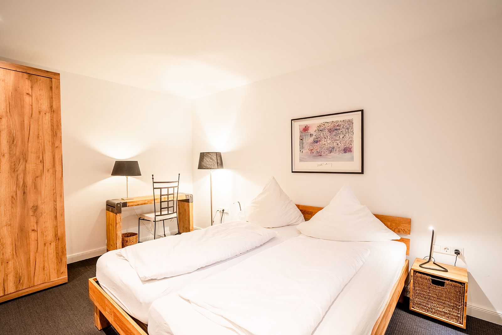 Helles Schlafzimmer mit Doppelbett, Holzschrank, Schreibtisch, zwei Lampen und Wandbild.