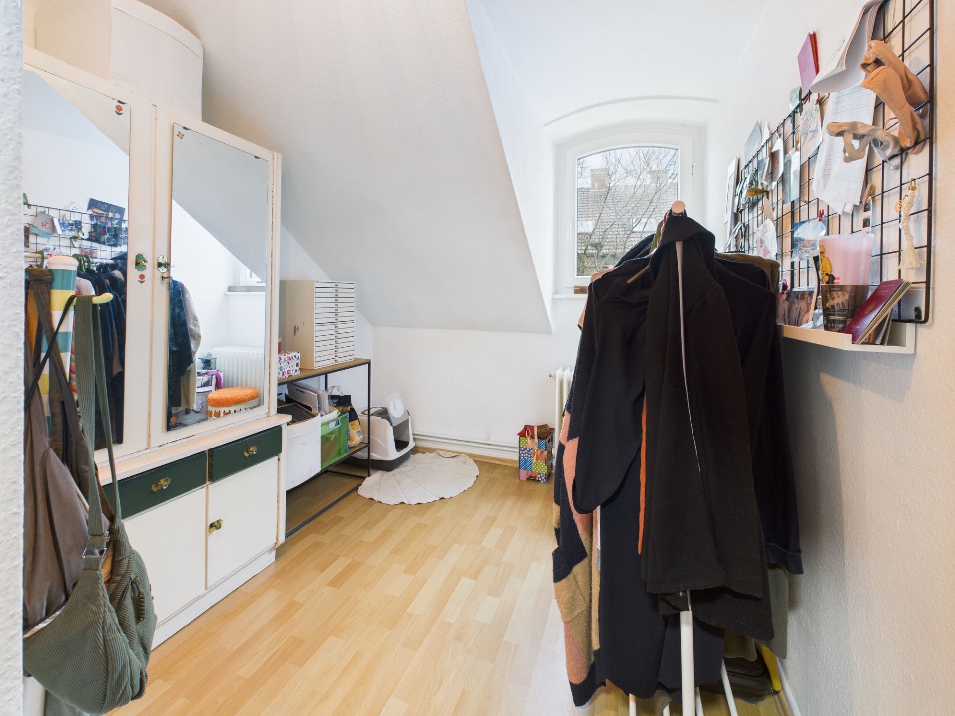 Helles Zimmer mit Dachschräge, Holzfußboden, Kleiderschrank, Spiegel und Fenster.