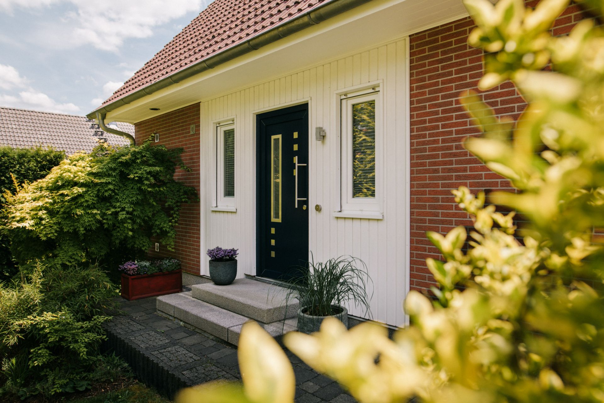 Eingang eines Einfamilienhauses mit dunkler Tür, zwei Fenstern und Pflanzen vor dem Haus.