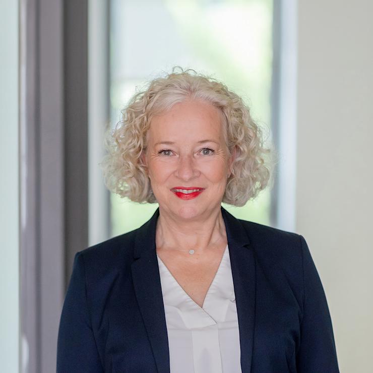 Eine ältere Frau mit lockigem, grauem Haar trägt einen dunklen Blazer und steht in einem hellen Raum.