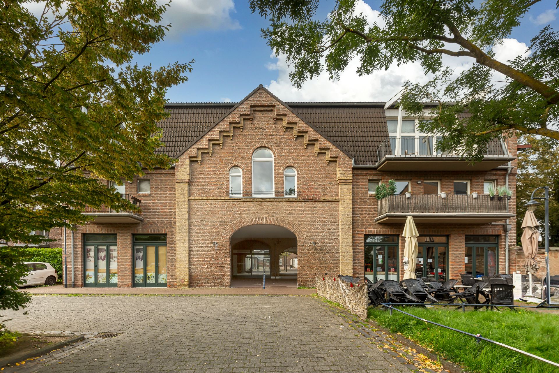 Backsteingebäude mit Balkon und großen Fenstern, zentrale Durchfahrt, davor Sitzgruppe und Bäume.