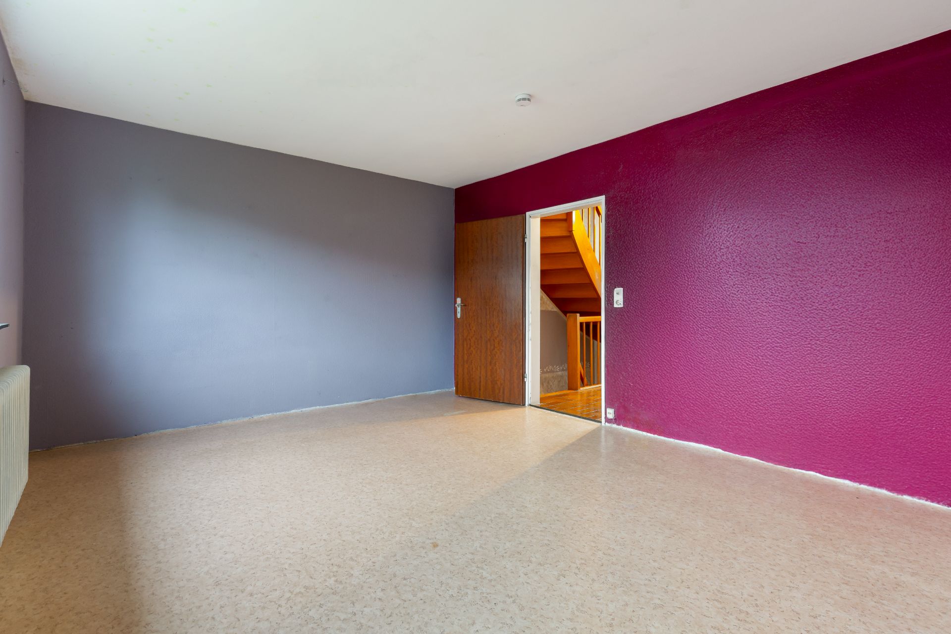 Leerer Raum mit lila Wand, grauer Wand, offener Tür und sichtbarer Holztreppe.