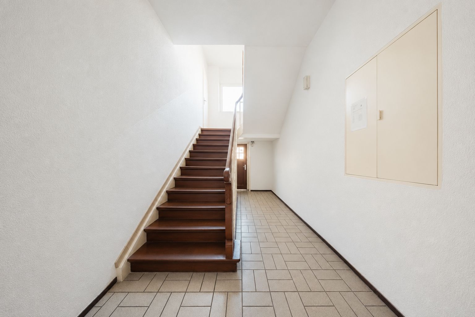 Heller Flur mit brauner Treppe und gefliestem Boden in einem Wohnhaus.
