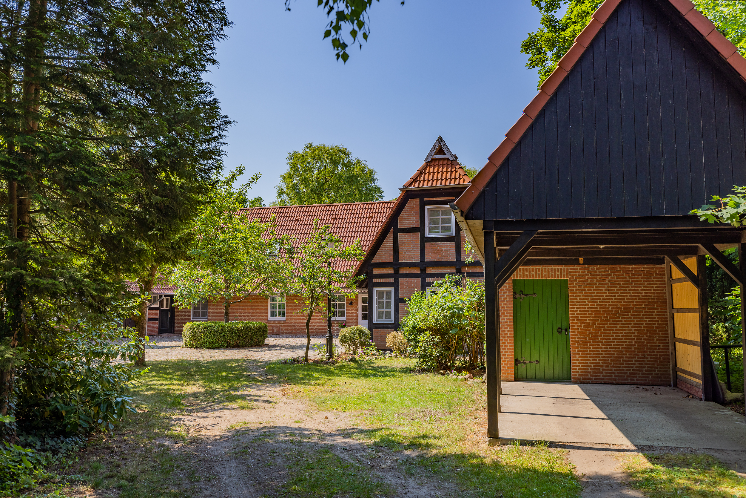 Traditionelles Backsteinhaus mit rotem Dach und Carport, umgeben von Bäumen und Garten.