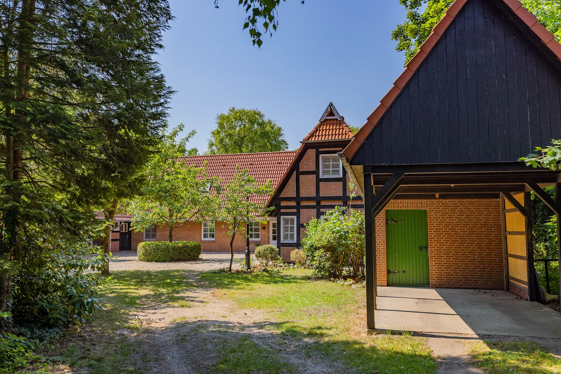 Traditionelles Backsteinhaus mit rotem Dach und Carport, umgeben von Bäumen und Garten.