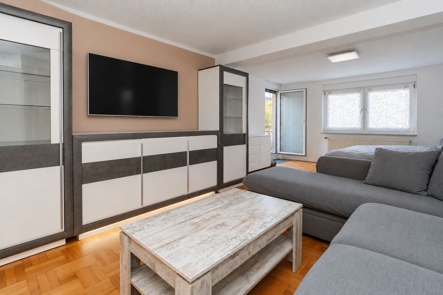 Modernes Wohnzimmer mit grauem Sofa, Couchtisch, TV an der Wand und Glastüren zu einem Balkon.