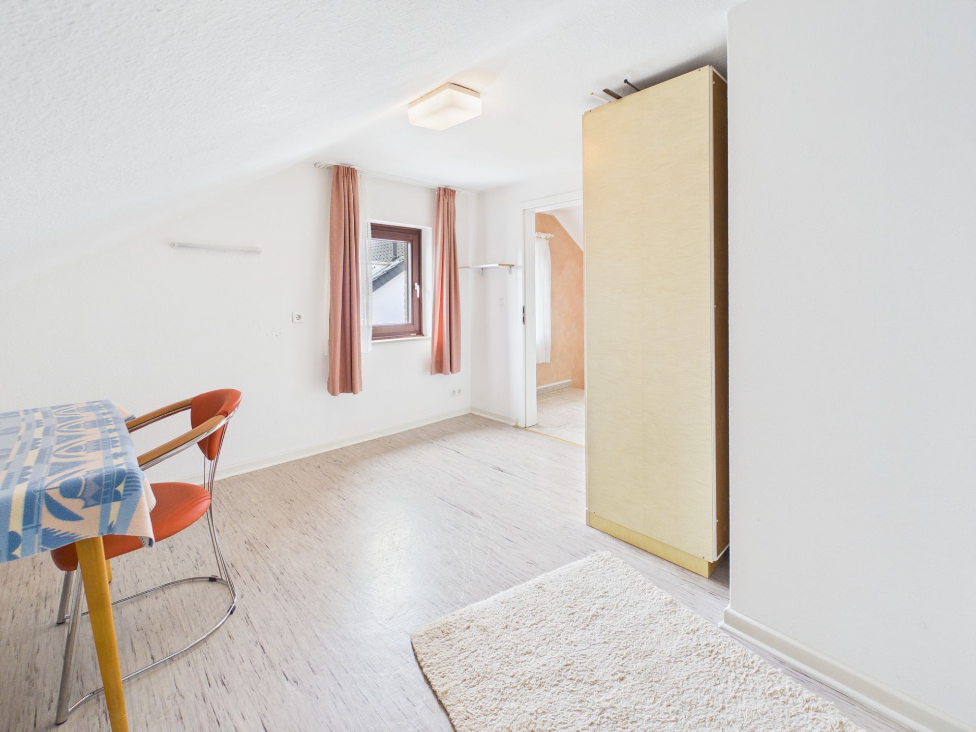 Helles Zimmer mit einem Fenster, Vorhängen, Tisch, Stuhl und Schrank.