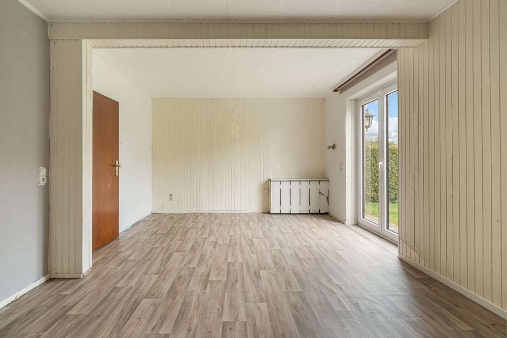 Leeres Zimmer mit Holzboden, weißer Wandvertäfelung und Glastür zum Garten.