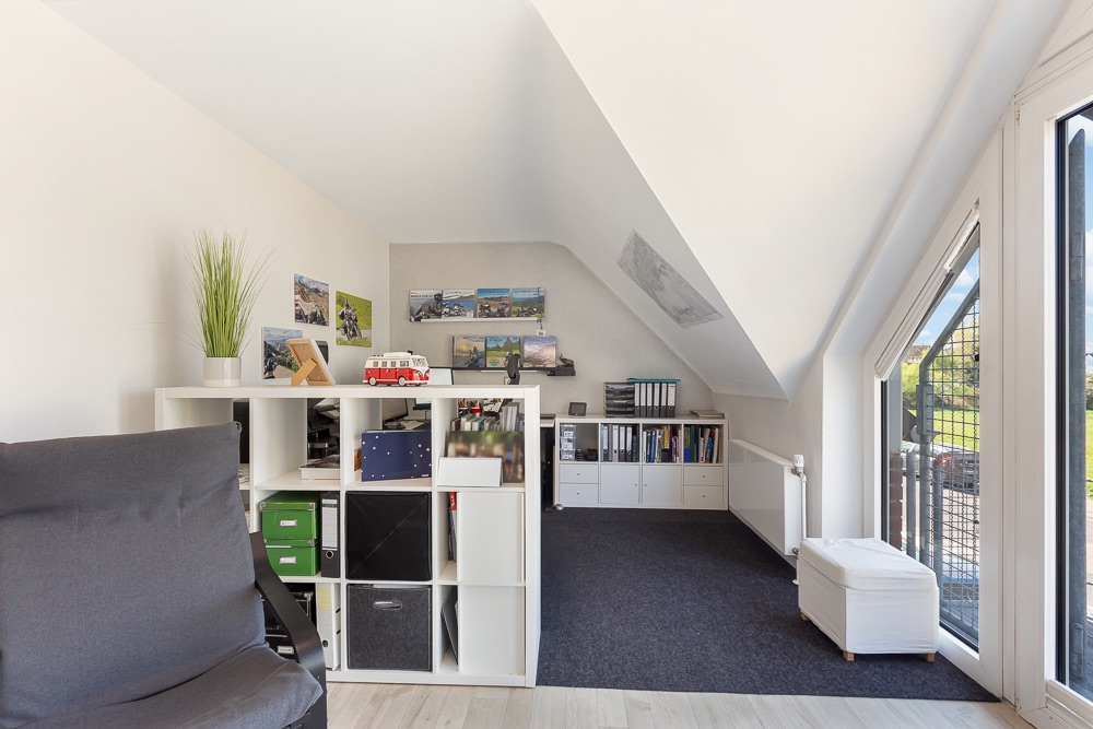 Helles Dachzimmer mit Regal, Sessel, Schreibtisch, Ordnern und großen Fenstern.
