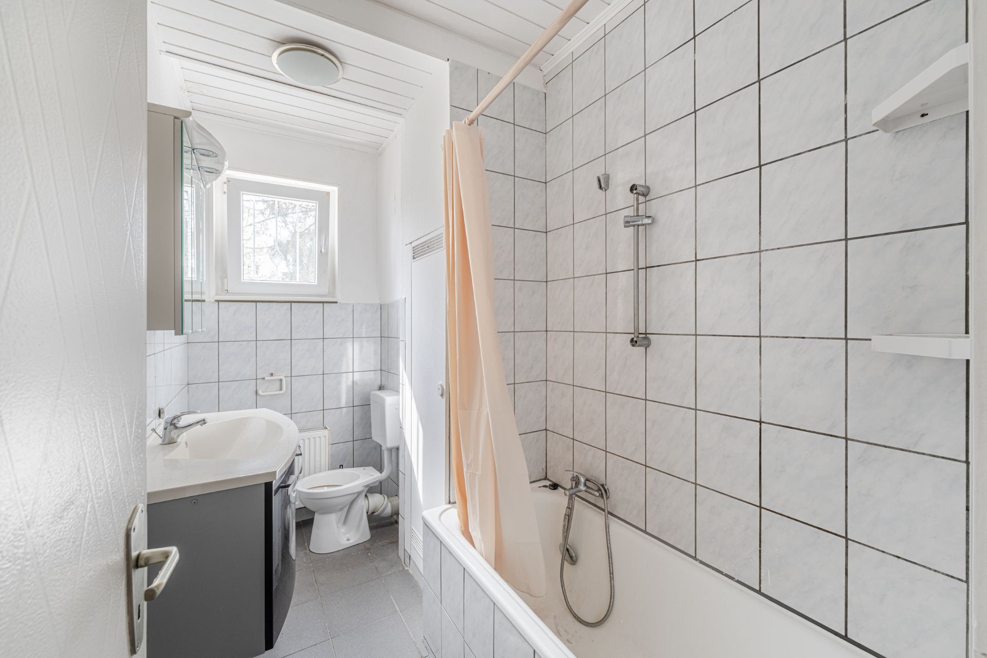 Helles Badezimmer mit Badewanne, Duschvorhang, WC und Waschbecken, grau gefliest.