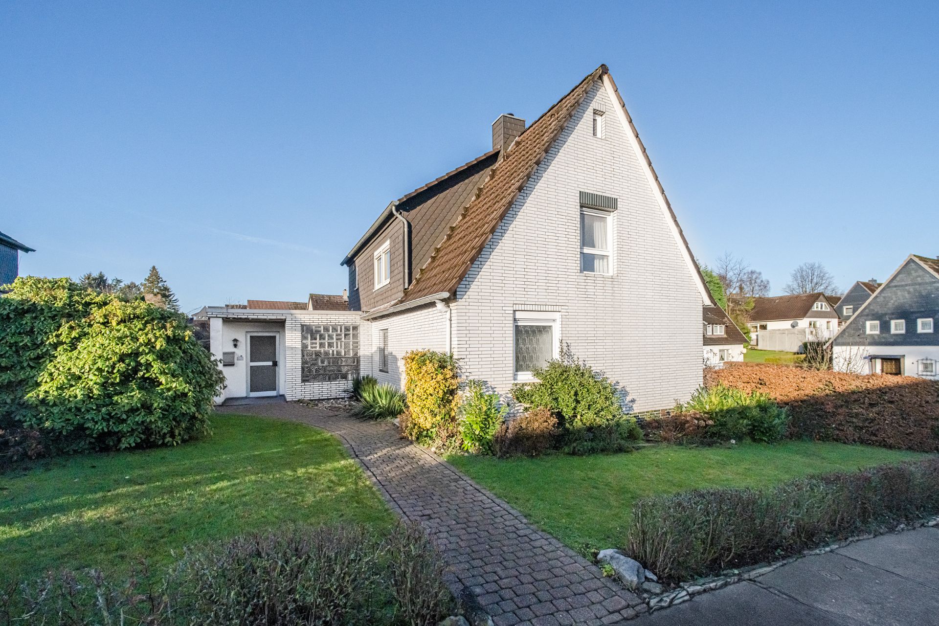 Weißes Einfamilienhaus mit Satteldach, Vorgarten und Gehweg bei klarem Himmel.