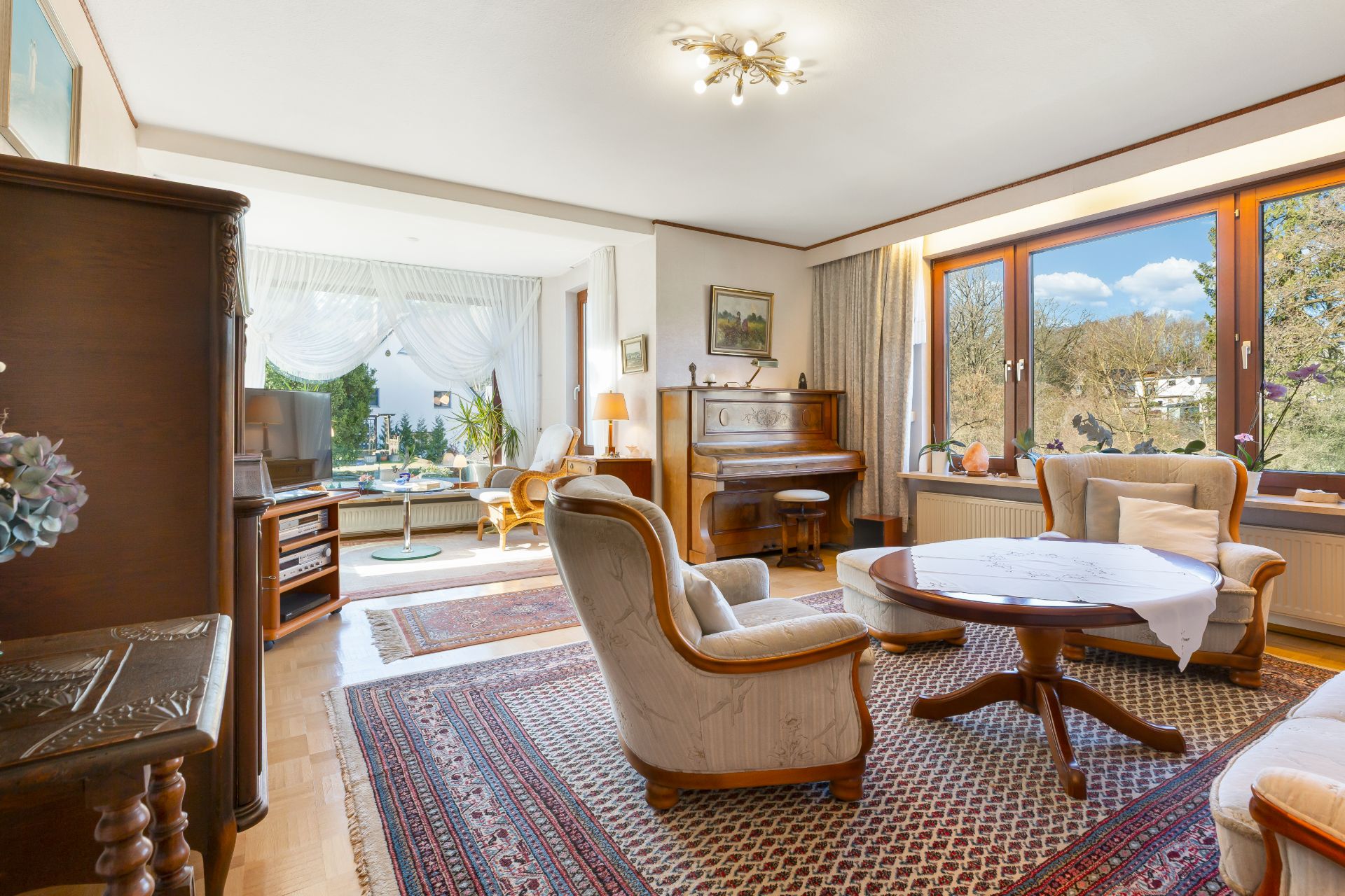 Wohnzimmer mit Teppich, Sesseln, Tisch, Klavier und großen Fenstern mit Blick ins Grüne.