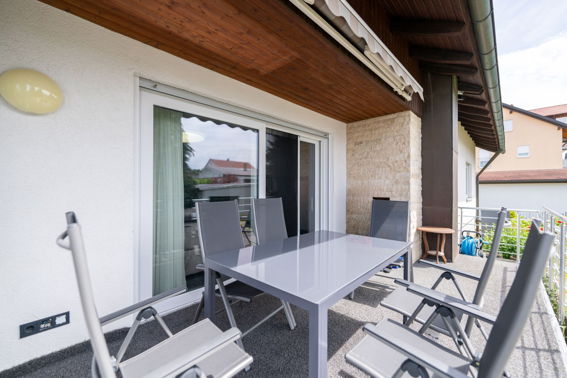 Moderne Terrasse mit Glastisch und sechs Stühlen vor einer weißen Hauswand und großen Fenstern.