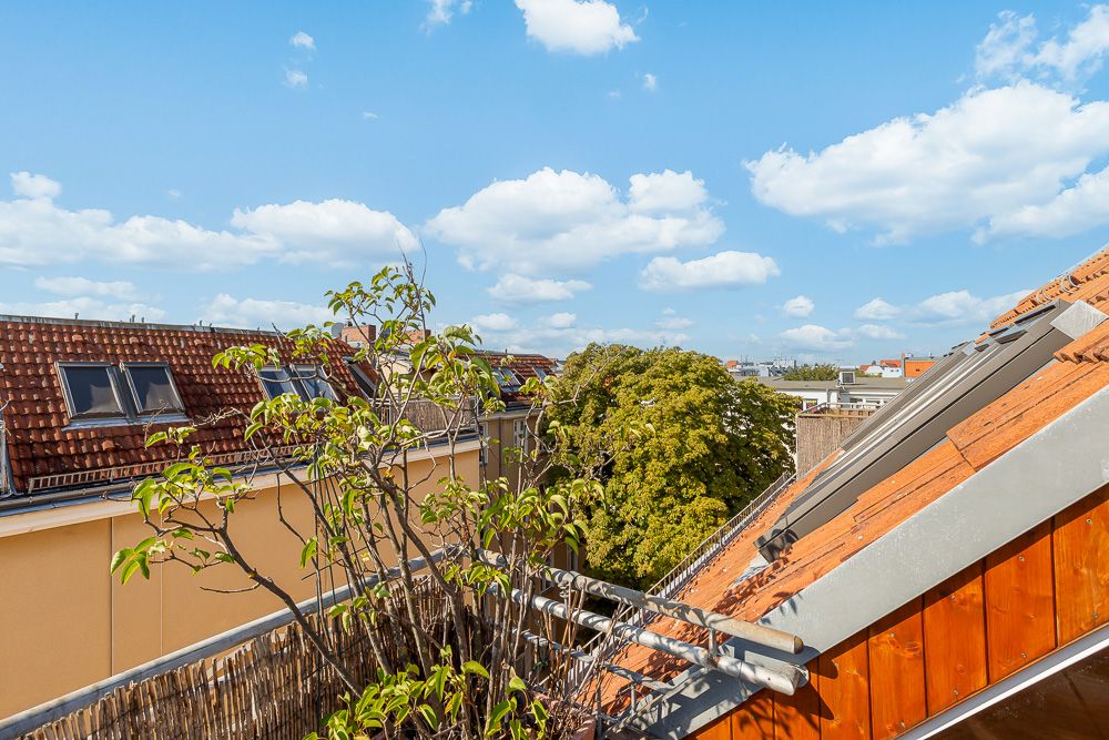 Blick von einem Balkon auf Dächer mit Dachfenstern und grüne Bäume unter blauem Himmel.