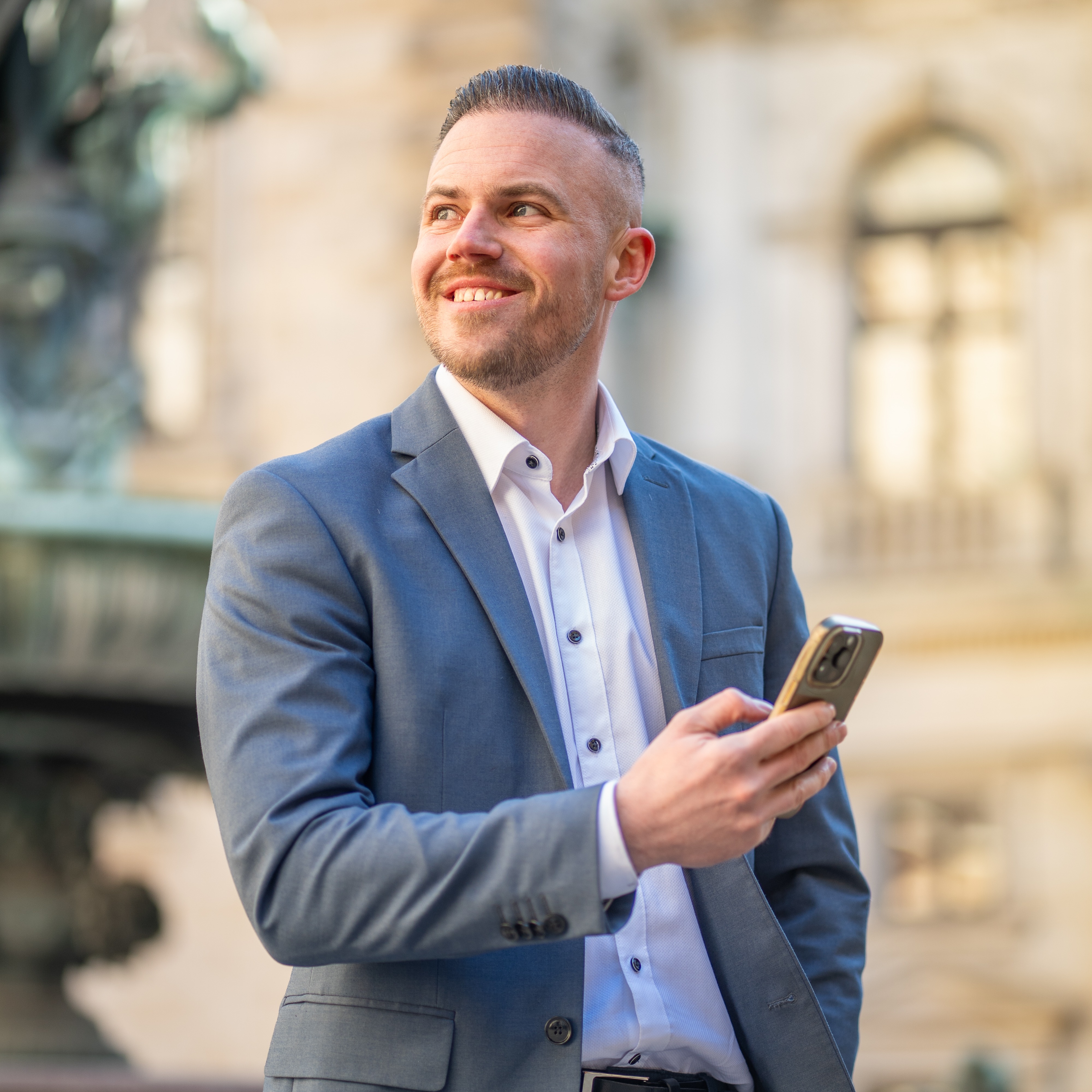 Ein Mann im Anzug hält ein Smartphone und lächelt im Freien.