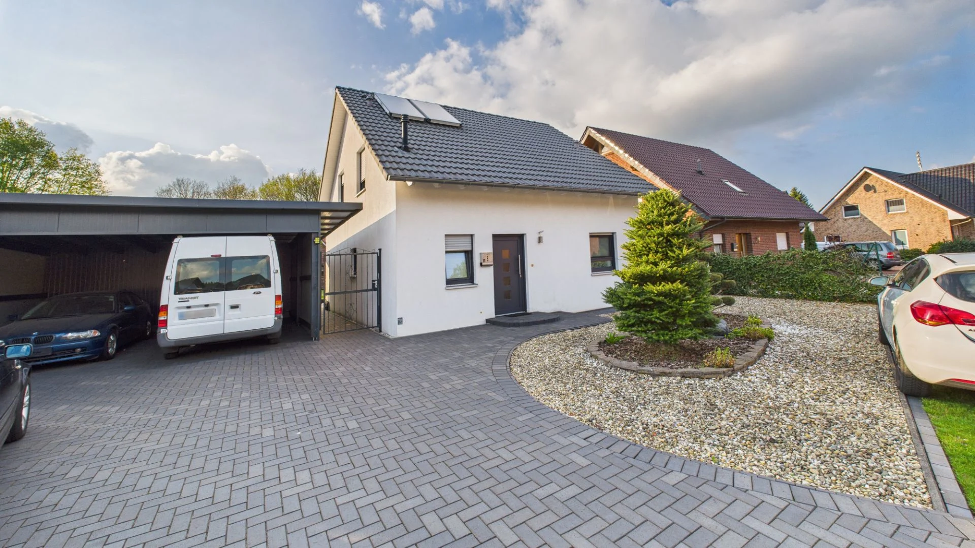 Modernes Einfamilienhaus mit Carport, gepflasterter Einfahrt und Kiesfläche im Vorgarten.
