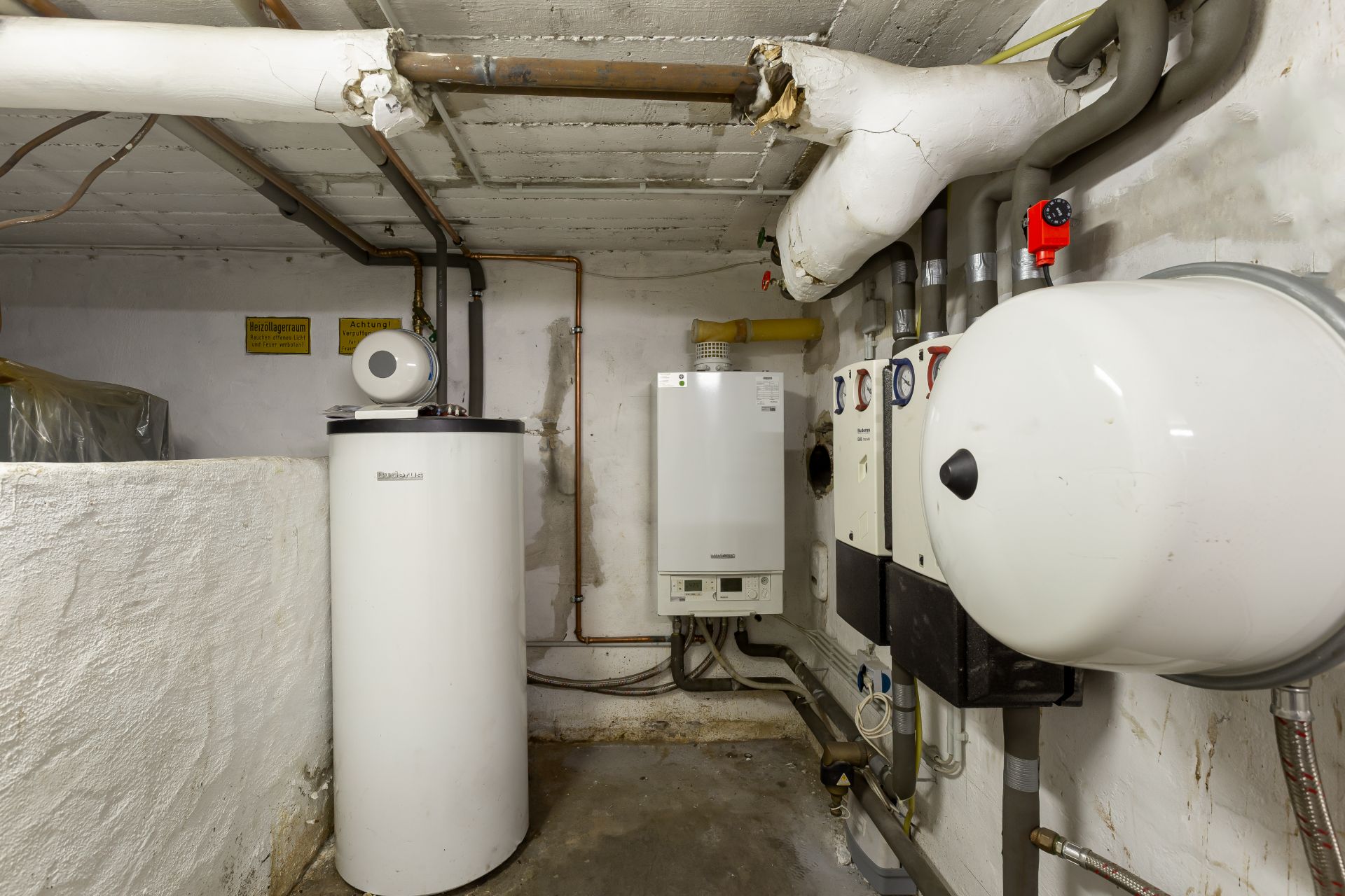 Heizungsraum mit Boiler, Rohren und Wandgeräten in einem Keller.