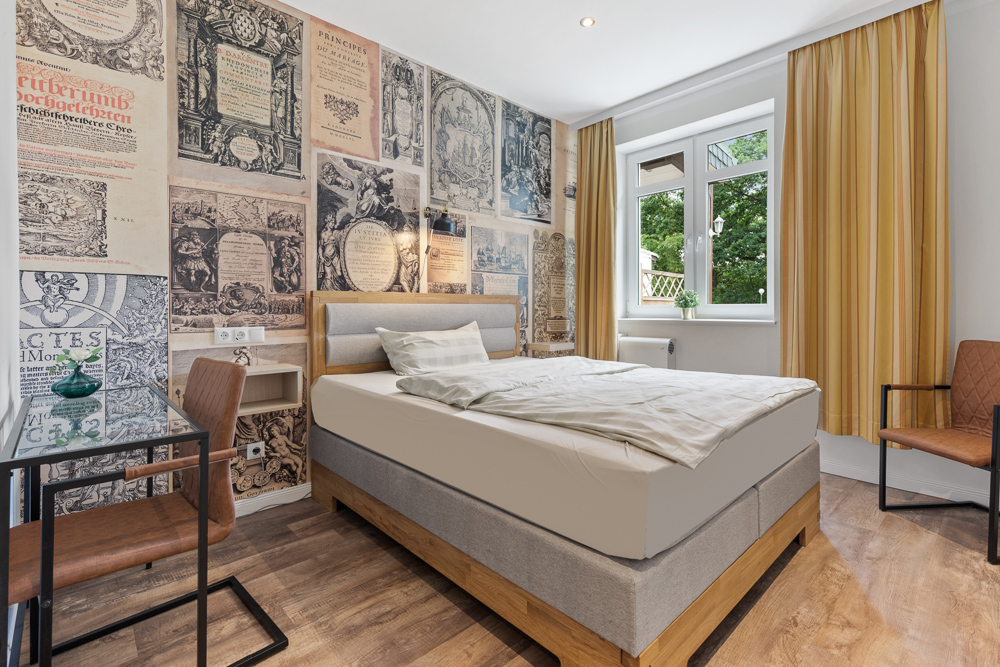 Helles Schlafzimmer mit Bett, Schreibtisch, zwei Stühlen und Wand mit historischen Buchseiten.