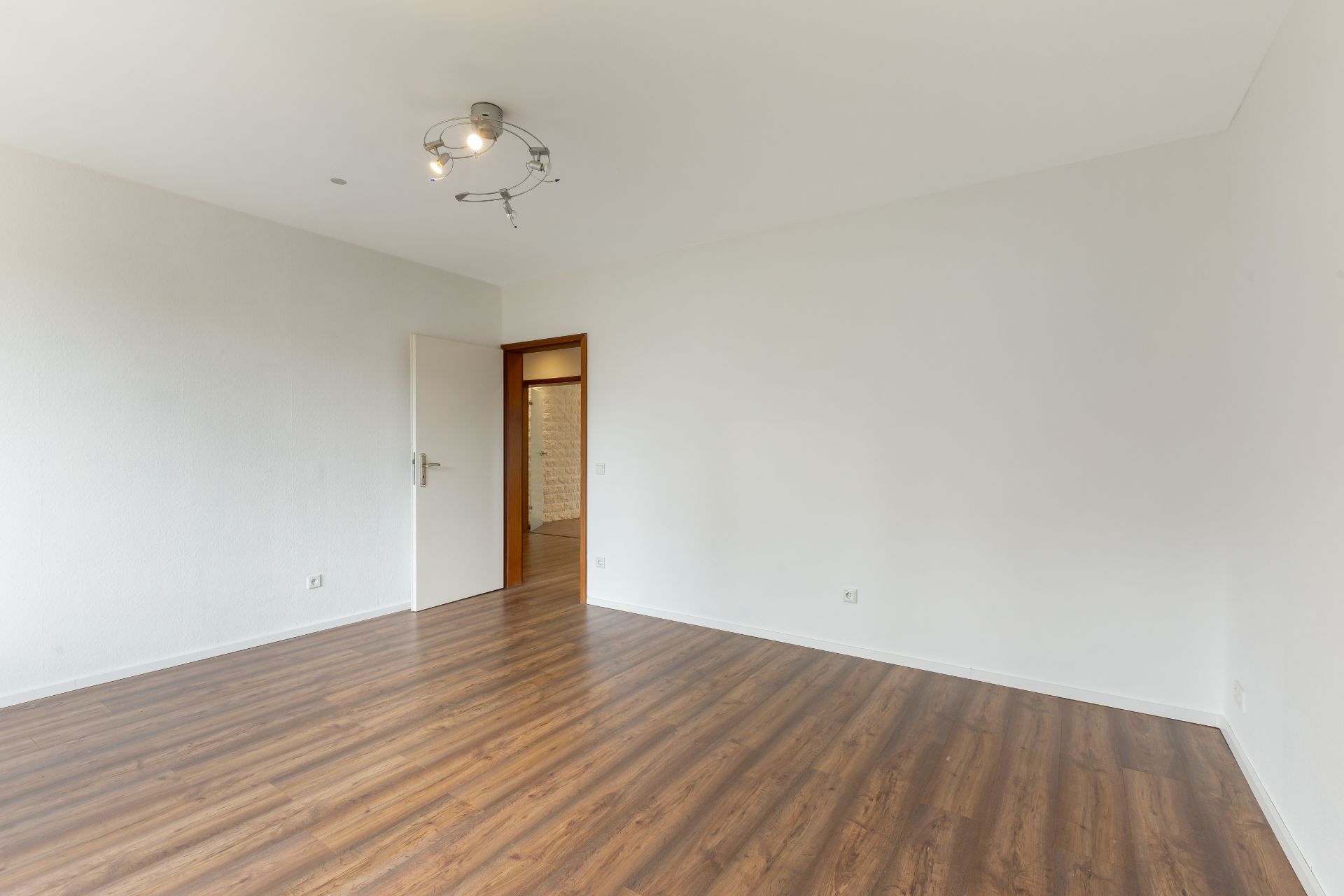Leeres Zimmer mit weißer Wand, Holzboden und Deckenlampe.