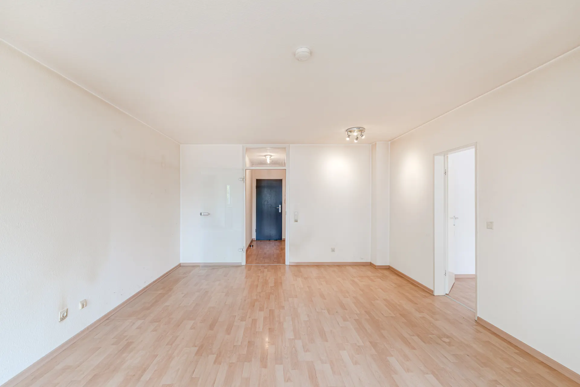 Leeres Wohnzimmer mit hellem Laminatboden und weißen Wänden, Türen führen in weitere Räume.